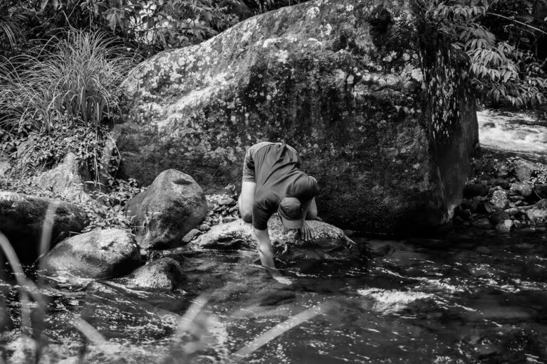 Black and white photograph of a man crouching on rocks at the edge of a fast-flowing river, one hand braced against a large mossy boulder, surrounded by dense vegetation.