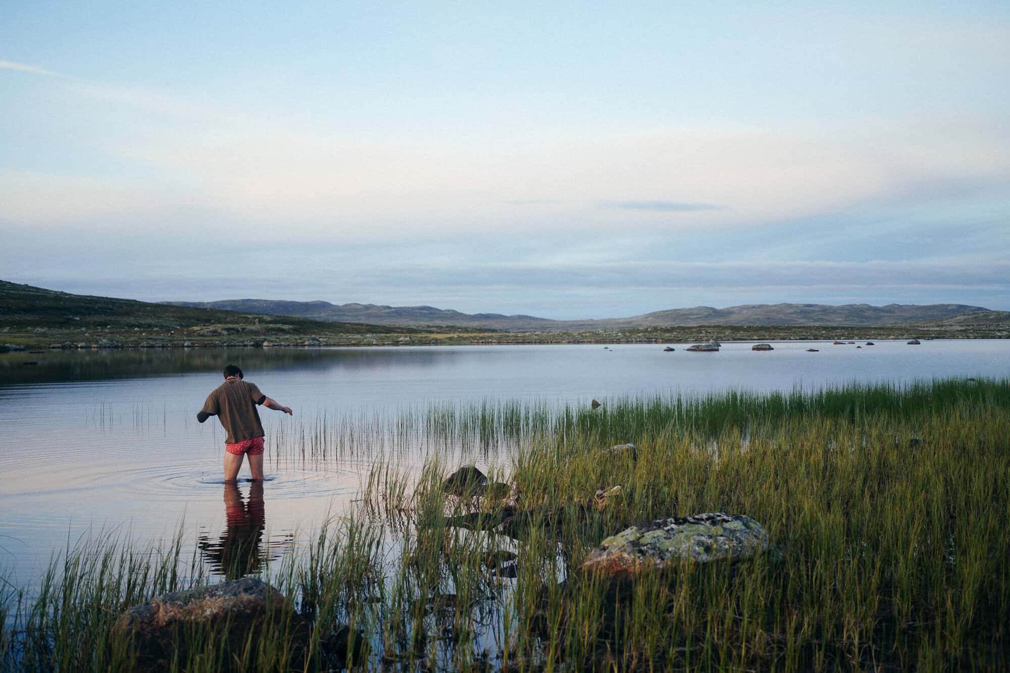 A man wades barefoot into a still lake at dusk, seen from behind, with reeds in the foreground and low hills on the far shore.
